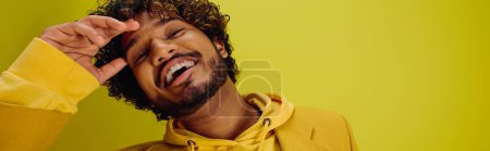 Ein gutaussehender junger indischer Mann in einem leuchtend gelben Kapuzenpulli macht vor einer eindrucksvollen Kulisse einen lustigen Gesichtsausdruck.