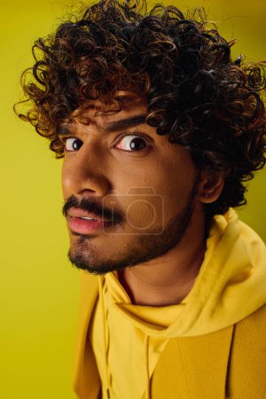 Handsome Indian man with curly hair posing in a vibrant yellow hoodie on a colorful backdrop.