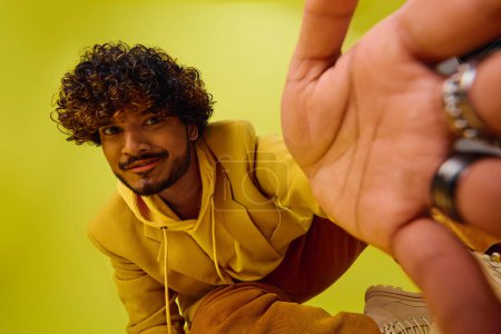 Hombre indio guapo con el pelo rizado posando en una sudadera con capucha amarilla sobre un fondo vívido.