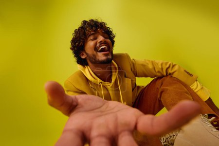 A handsome young Indian man poses in a bright yellow hoodie, extending his hand outwards.