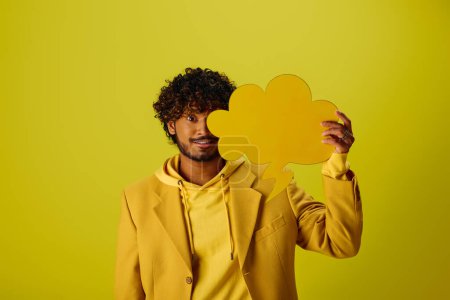 Hombre indio guapo en chaqueta amarilla sosteniendo la burbuja del habla contra el telón de fondo vibrante.