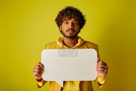 Ein junger indischer Mann in einem gelben Kapuzenpullover hält eine Sprechblase vor einem lebhaften Hintergrund.