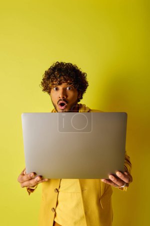 A man in vibrant clothing obscures his face with a laptop.