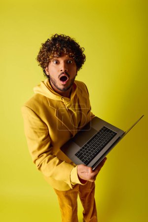 Photo for Handsome young Indian man with curly hair holding a laptop on vibrant backdrop. - Royalty Free Image