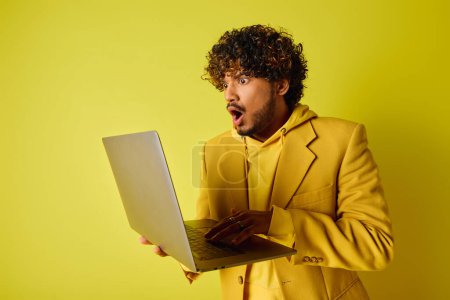A stylish young Indian man in a yellow suit confidently holding a laptop.