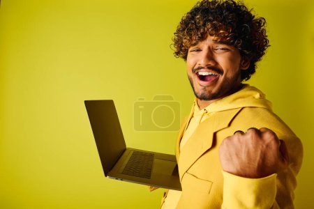 Téléchargez les photos : Beau jeune Indien en costume jaune vibrant tient avec confiance un ordinateur portable. - en image libre de droit