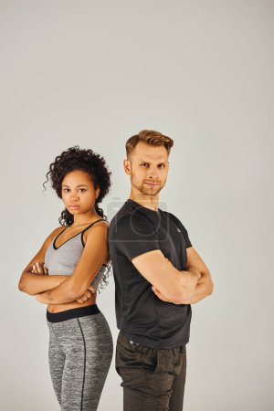 Ein junges interrassisches Sportpaar in aktiver Kleidung posiert selbstbewusst vor grauem Studiohintergrund.
