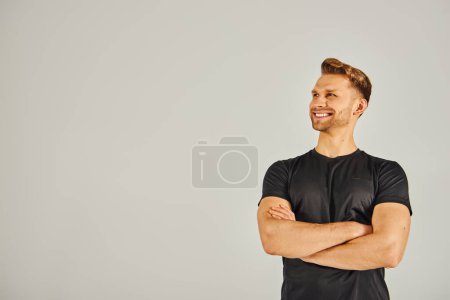 Foto de Un joven atlético con una camiseta negra posa confiadamente con los brazos cruzados en un estudio sobre un fondo gris. - Imagen libre de derechos