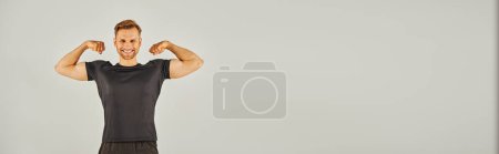 Junger, athletischer Mann in aktiver Kleidung, der seinen Bizeps vor grauem Hintergrund in einem Studio-Setting spannt.