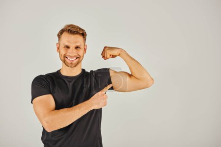 Photo for A young athletic man in active wear flexing his biceps against a gray background, showcasing strength and determination. - Royalty Free Image