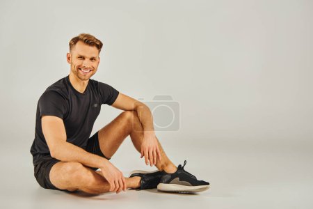 Un joven atlético en ropa activa se sienta en el suelo, en el fondo del pensamiento, sus zapatillas visibles.