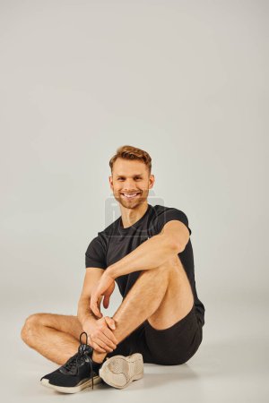 Foto de Un joven atlético en ropa activa sentado en el suelo y sonriendo alegremente en un estudio con un fondo gris. - Imagen libre de derechos