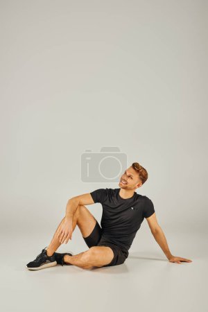 Téléchargez les photos : Un jeune homme athlétique assis sur le sol dans un t-shirt noir, perdu dans la pensée, dans un studio avec un fond gris. - en image libre de droit