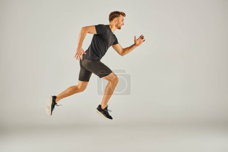 A young athletic man in active wear is energetically running on a grey background in a studio setting.