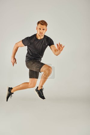 A young athletic man in a black t-shirt is energetically jumping in a studio with a grey background.