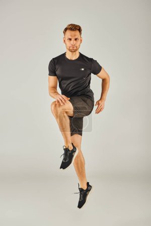 A young athletic man in a black t-shirt and shorts jumps energetically on a grey background in a studio setting.