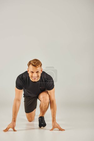 Ein junger, athletischer Mann in aktiver Kleidung kauert vor grauem Hintergrund in einem Studio-Setting.