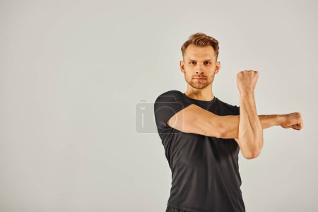 Foto de Un joven atlético en ropa activa flexiona su brazo contra un fondo gris en una dinámica muestra de fuerza y forma física. - Imagen libre de derechos