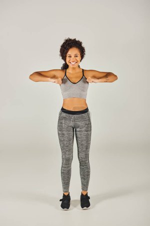 Foto de Mujer afroamericana joven y en forma que golpea una pose elegante con los brazos extendidos en desgaste activo en el fondo gris del estudio. - Imagen libre de derechos