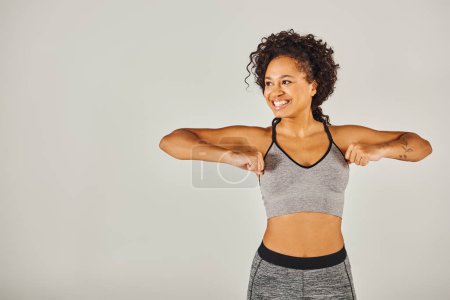 Foto de Una joven afroamericana en ropa deportiva enérgicamente flexiona sus brazos frente a un fondo gris. - Imagen libre de derechos