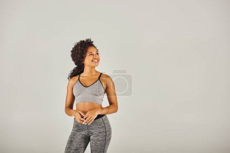 Téléchargez les photos : Une jeune femme afro-américaine en soutien-gorge de sport et leggings faisant une routine d'entraînement dans un studio sur fond gris. - en image libre de droit