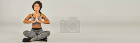 Young African American woman in activewear gracefully practicing yoga on a serene gray background in a studio setting.