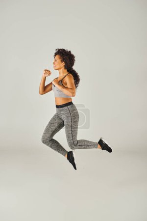 A young African American woman performs a dynamic jump in a grey sports bra and leggings in a studio setting.