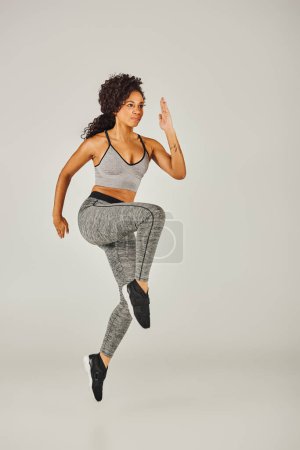 Una joven afroamericana en forma en sujetador deportivo y polainas salta poderosamente sobre un fondo gris de estudio.