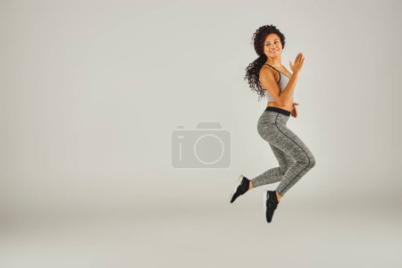 Una joven afroamericana vestida con atuendo atlético salta alegremente sobre un fondo gris en un ambiente de estudio.