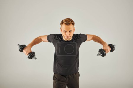 Foto de Un joven deportista en ropa activa sostiene dos pesas en un estudio con un fondo gris. - Imagen libre de derechos