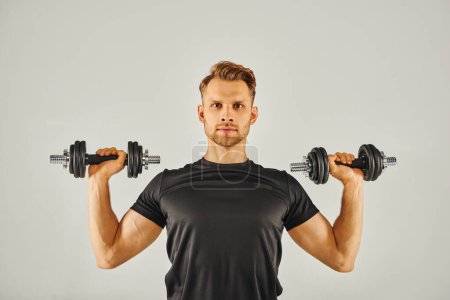 Ein junger Sportler in aktiver Kleidung hält in einem Studio vor grauem Hintergrund zwei Kurzhanteln, die sich auf Fitness und Kraft konzentrieren.