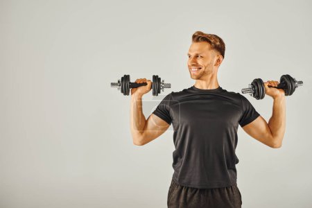 Photo for A young sportsman in active wear holds two dumbbells in front of a gray background in a fitness studio. - Royalty Free Image