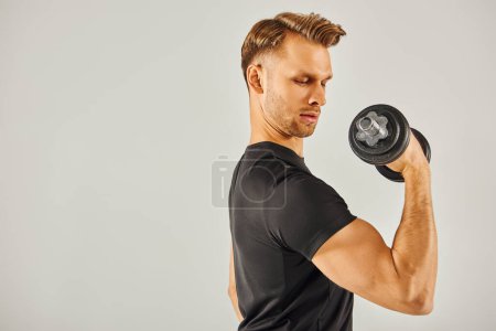 Photo for A young sportsman in active wear lifts a dumbbell in a studio with a grey background. - Royalty Free Image