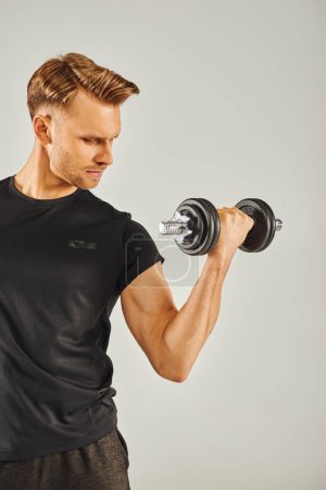 Ein junger Sportler in aktiver Kleidung demonstriert seine Stärke, indem er in einem Studio mit grauem Hintergrund ein Paar Kurzhanteln hält..