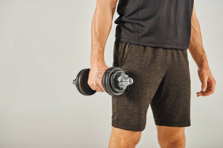 A young sportsman in active wear energetically lifts a pair of dumbbells in a studio with a grey background.