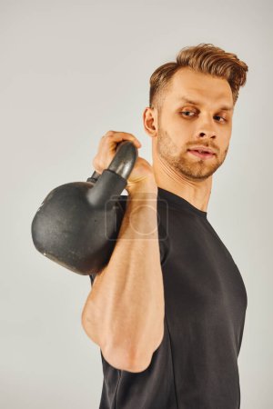 A young sportsman in active wear showcasing his strength by holding a kettlebell on his arm against a grey studio background.