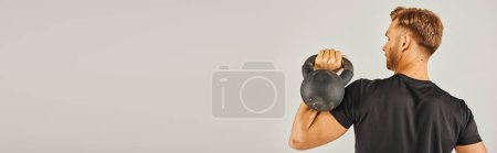 Photo for A young sportsman in active wear lifts a kettlebell with determination in a studio setting with a grey background. - Royalty Free Image