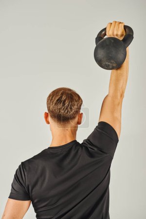 Un joven deportista en ropa activa levanta intensamente una campana de agua en un estudio con un fondo gris.