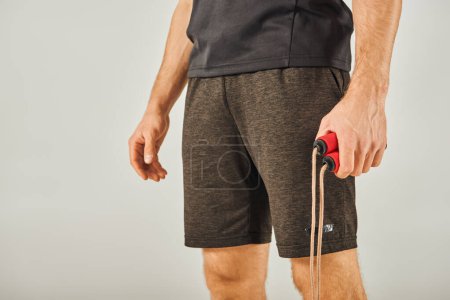Téléchargez les photos : A young sportsman in activewear is skillfully holding a vibrant red and black skipping rope in a studio with a grey background. - en image libre de droit