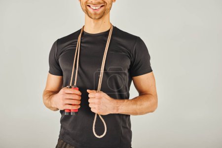 Foto de Un joven deportista en ropa deportiva sonríe mientras sostiene una cuerda saltando en un estudio con un fondo gris. - Imagen libre de derechos