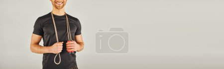 Téléchargez les photos : A young sportsman in active wear smiling while holding a rope in a studio with a grey background. - en image libre de droit