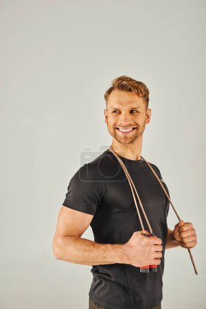 Foto de Joven deportista en activo sonriendo mientras sostiene una cuerda saltando en un estudio con un fondo gris. - Imagen libre de derechos
