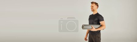 Photo for Young sportsman in active wear holding a yoga mat in front of a grey background. - Royalty Free Image