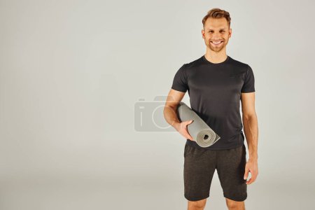A young man in active wear holding a yoga mat in front of a gray background.