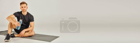 Photo for A young sportsman in active wear focuses on perfecting his squat position while sitting on a mat in a studio with a grey background. - Royalty Free Image
