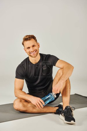 Ein junger Sportler in aktiver Kleidung sitzt auf einer Yogamatte, hält eine Wasserflasche in der Hand und macht nach einer Sitzung eine Pause..
