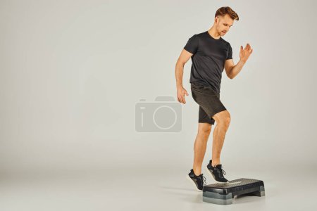 Un joven deportista en ropa activa utiliza un paso en un estudio sobre un fondo gris.