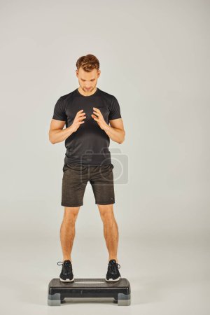 Un jeune sportif en tenue active remet en question son équilibre sur un stepper dans un studio au fond gris.