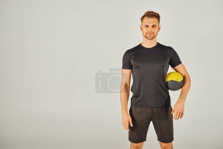 Joven deportista en camiseta negra demostrando ejercicio físico con una vibrante bola amarilla y negra en un ambiente de estudio.