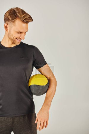 Junger Sportler im schwarzen T-Shirt mit gelbem Ball in einem Studio mit grauem Hintergrund.